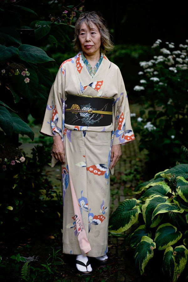 colour phootgraph of japanese woman in  her sixties dressed in a traditional japanese beige kimono with a pattern of red blue, white and black coloured butterflies. She is standing in a shady part of a garden with white flowers on a shrubbery, a multi-coloured host in the foreground and small pink flowers to her left. Her gaze is turned away from the lens, she looks contented and peaceful.