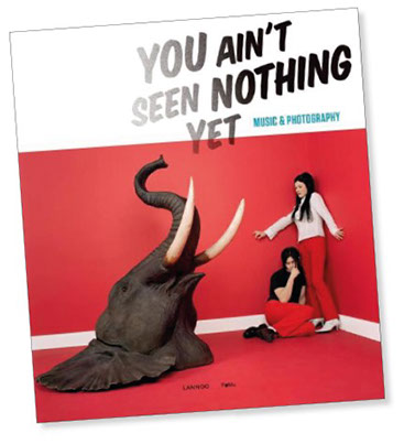Tilted colour photograph of book cover with the title in black You Ain't seen nothing yet and the subtitle in blue: Music and Photography. who thirds of the book cover contains a corner of room with red walls and red floor and a young man seated on the ground an a young woman standing next to him in the corner trying to get away from the giant head of an elephant on the floor. The people wear red tight trousers, the girl wears a white shirt with long sleeves, the man a black T shirt, both look in agony to the elephant.