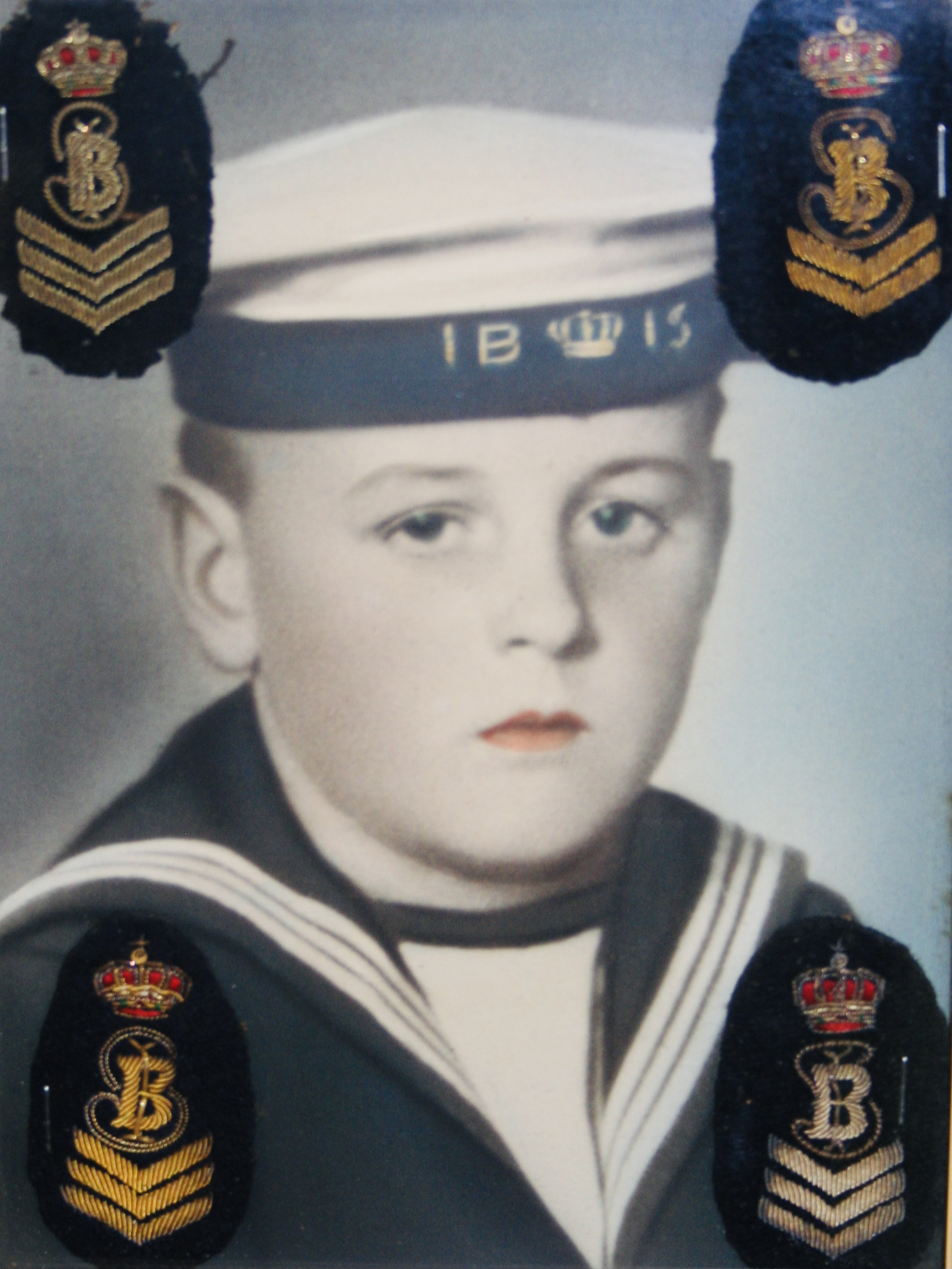 a hand-coloured black and white identity portrait of a boy in a sailor suit with blue eyes and orange lips. On his sailor hat it says IBIS  and the hat also has a crown, four emblems are sewn in  the corners of the photograph