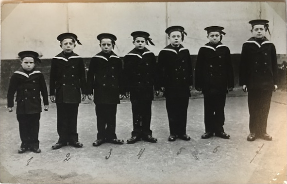 a black and white photograph of seven young boys in dark sailor suits. The y stand in a line, with the youngest, who looks tiny like a six year old on the far left. The other six boys look bigger and older.