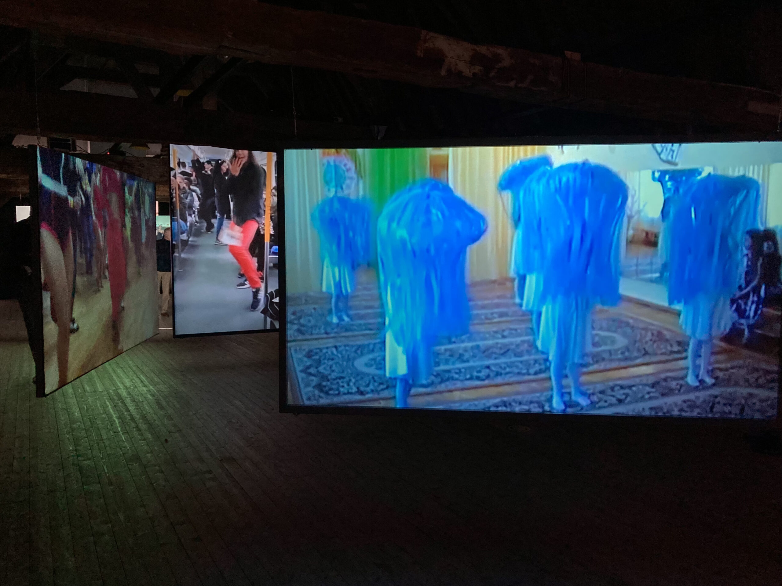 A colour photograph of a darkened exhibition space with three screens,, each showing a different video of dancing people  at various locations. the largest screen shows six dancing people wearing blue costumes  which makes them look like sea anemones